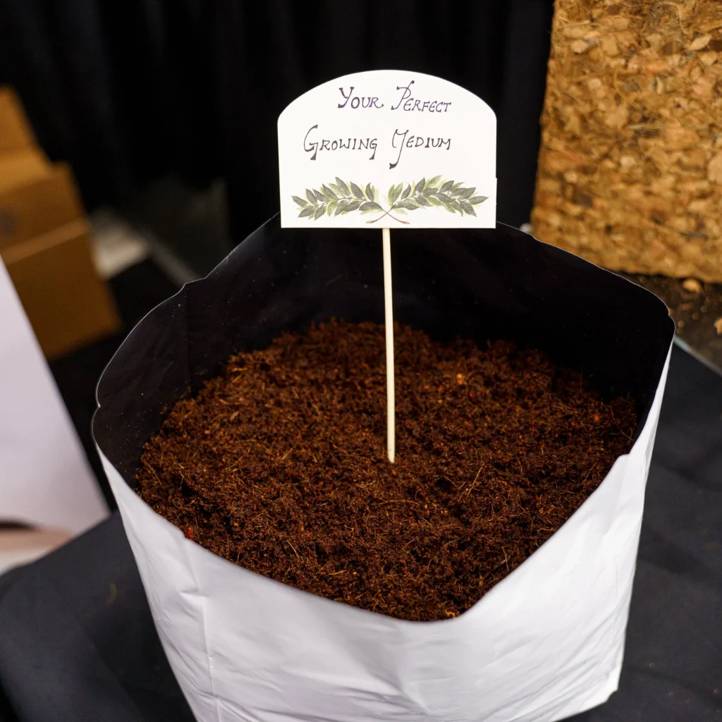 A white, open-top grow bag filled with dark, hydrated coco coir growing medium, with a small sign stick reading "Your Perfect Growing Medium" inserted into the soil.
